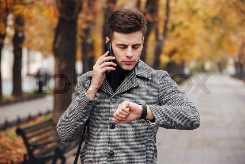 Picture of businesslike man speaking on mobile phone while going on meeting, checking time with watch on hand, stock photo