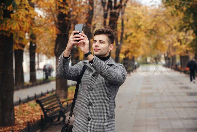 Handsome man in coat taking photo of beautiful autumn trees using his modern cellphone, while walking in empty park, stock photo