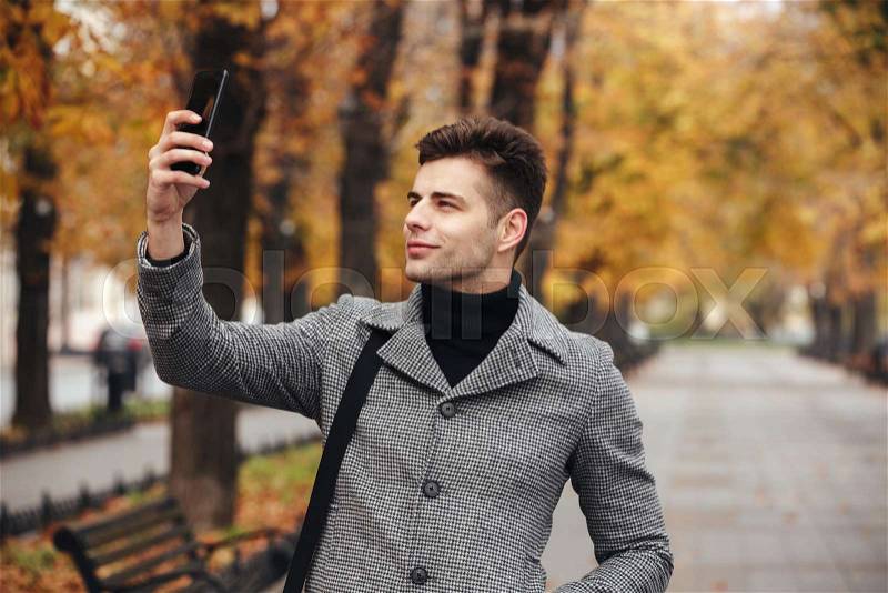 Cheerful man in coat taking photo of nature or making selfie using black smartphone, while walking along boulevard, stock photo