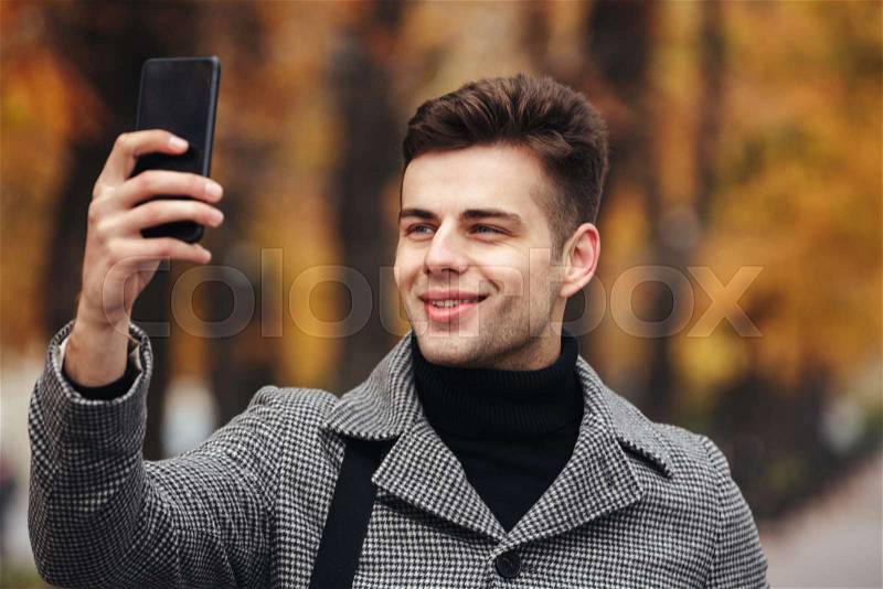 Happy man warmly dressed taking photo of nature or making selfie using black smartphone, while walking in park, stock photo