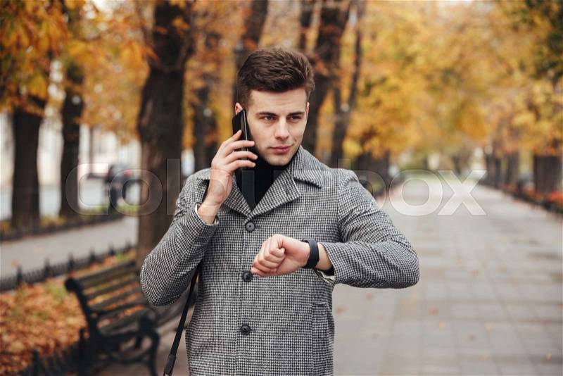 Picture of elegant man checking time with watch on hand, and speaking on mobile phone during his walk in park, stock photo