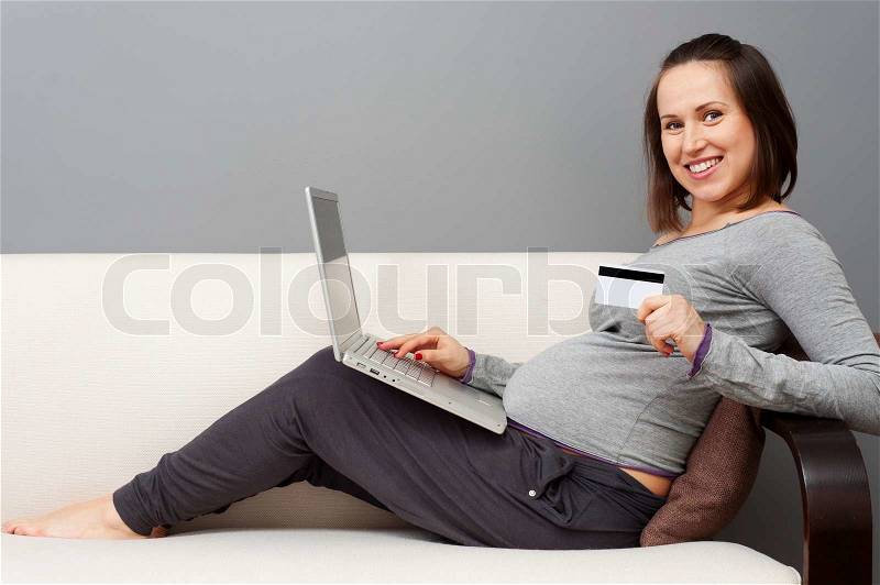 Smiley young pregnant woman holding laptop and showing credit card, stock photo