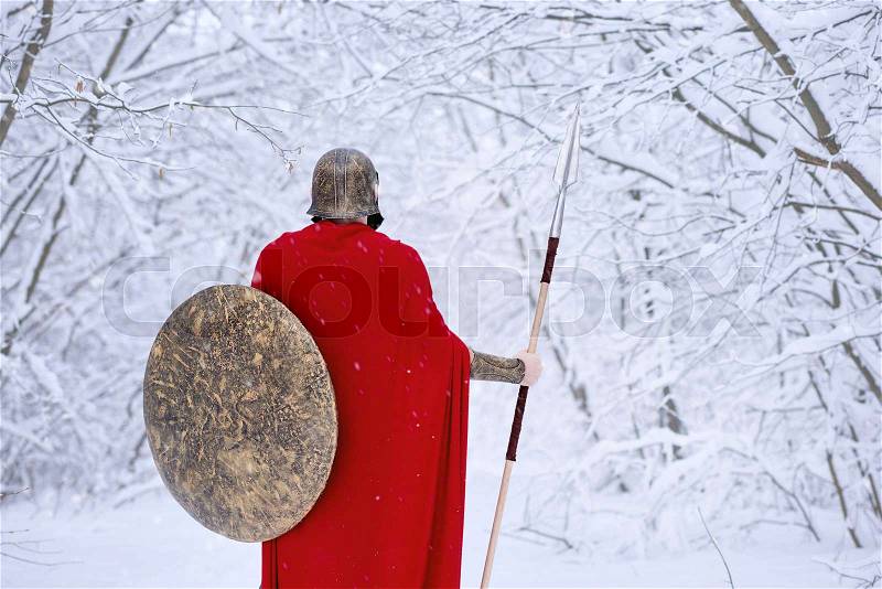 Careful spartan in cold snowy forest. A man looks brave and strong. He keeps traditional spartan armor: round big shield and sharp metallic spear,helmet on his head. Warrior wears long red cape, stock photo