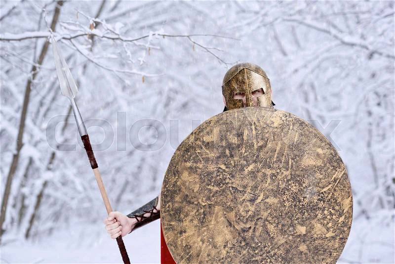 Spartan warrior wearing red long cape is looking in camera standing in white dense winter forest. He has metallic spear, shield and helmet on head. All trees covered with snow. It\'s cold, frosty, stock photo