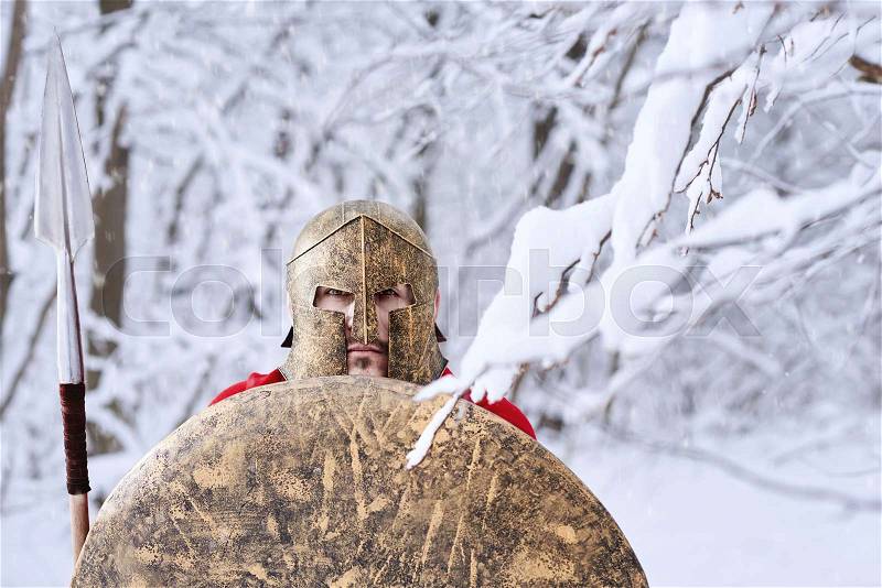 Spartan warrior wearing red cape is looking in camera standing in white dense winter forest. He has metallic spear, shield and helmet on head. All trees covered with snow. Man is strained, wary, stock photo