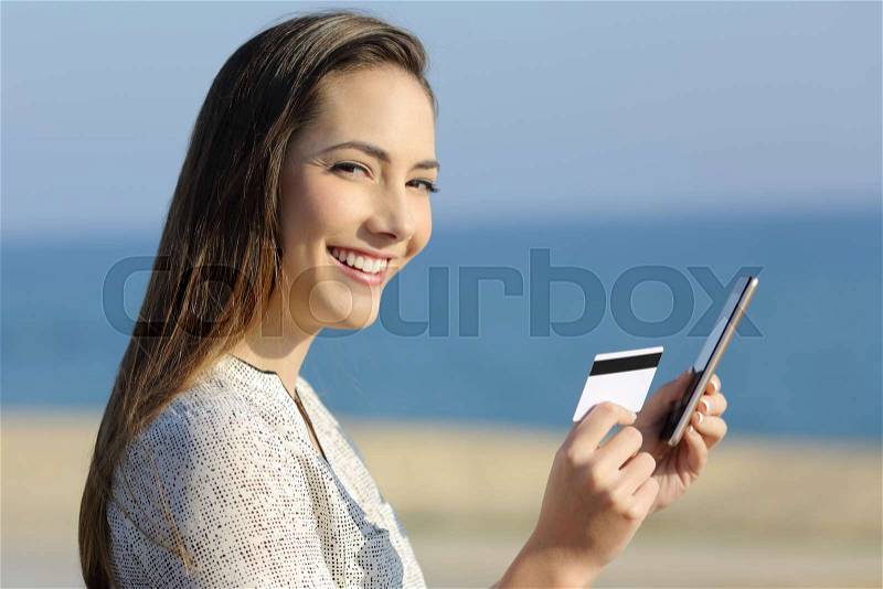 Happy girl holding a credit card and a smart phone looking at camera on the beach, stock photo