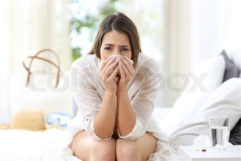 Front view portrait of an ill allergic woman looking at camera sitting on the bed of an hotel room on summer vacations, stock photo