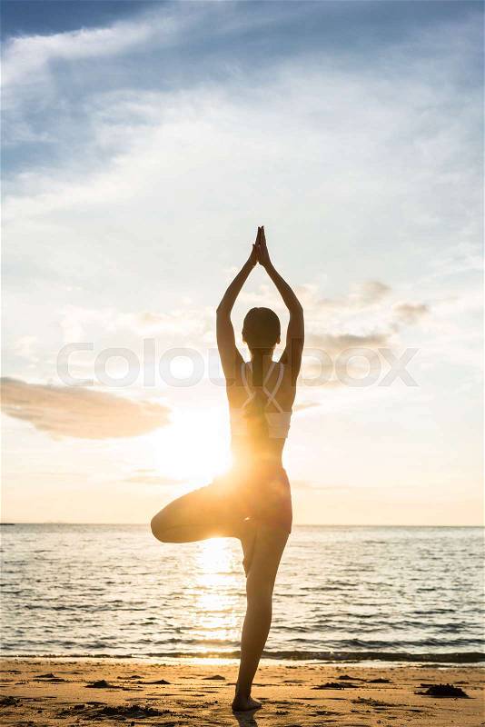 Full length rear view of the silhouette of a woman standing on one leg, while practicing the tree yoga pose on a tranquil beach at sunset during summer vacation in Indonesia, stock photo