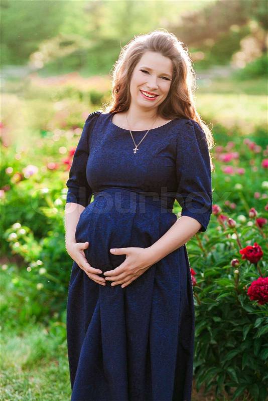 Pregnant woman in peony orchard holding tummy in heart form. Vertical shot, stock photo