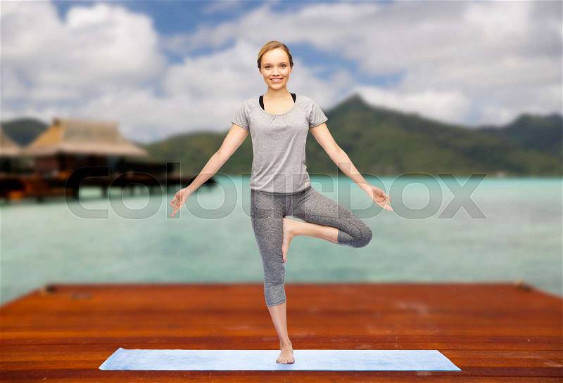 Fitness, sport, people and healthy lifestyle concept - woman making yoga in tree pose on wooden pier over island beach and bungalow background, stock photo