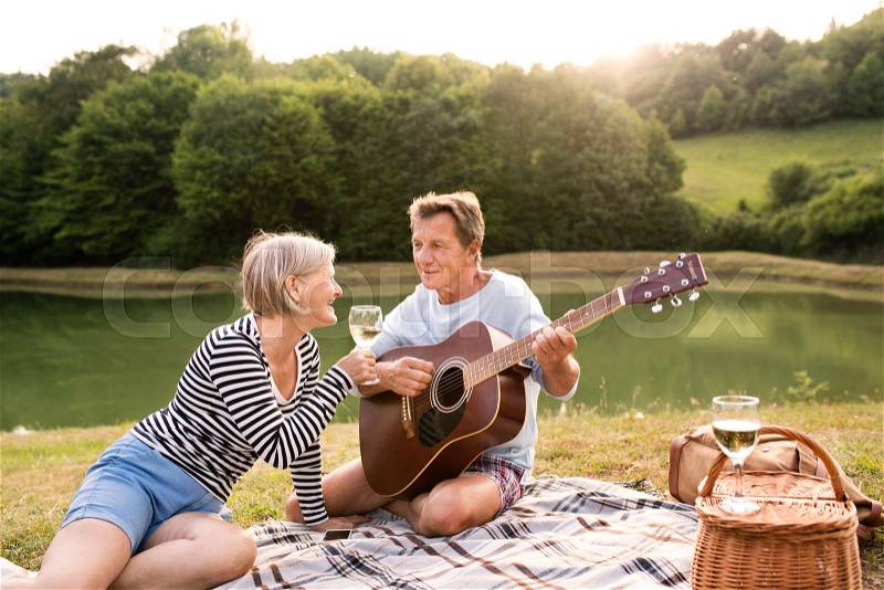 Beautiful senior couple at the lake having a picnic, sitting on blanket, drinking wine. Man playing guitar, stock photo