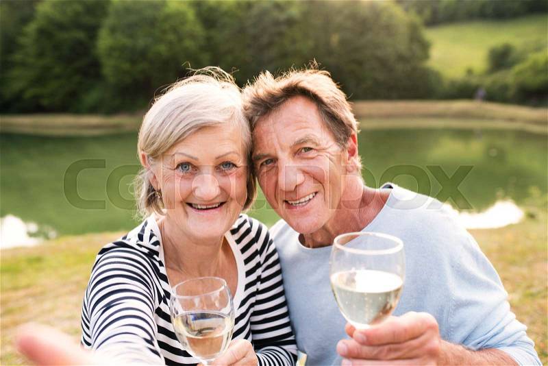Beautiful senior couple at the lake having a picnic, sitting on blanket, drinking wine, stock photo