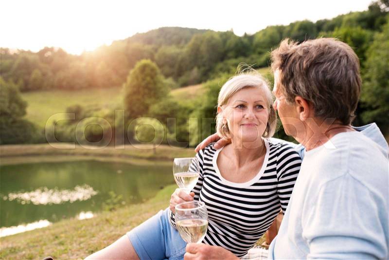 Beautiful senior couple at the lake having a picnic, sitting on blanket, drinking wine, stock photo