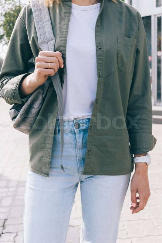 Details of everyday fashionable clothes. Close-up young woman wearing casual outfit: white top, khaki shirt and blue jeans holding a backpack standing on the street in the spring sunny day, stock photo