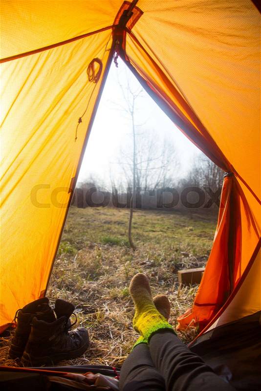 A person sitting in an orange tent, camp in the bank of the river in spring. Feet selfie of traveler. Relaxed, colorful look. Spring hiking, stock photo