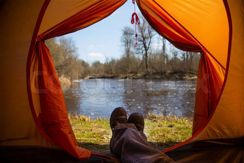A person sitting in an orange tent, camp in the bank of the river in spring. Feet selfie of traveler. Relaxed, colorful look. Spring hiking, stock photo