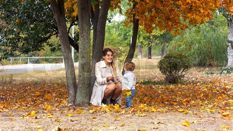 Happy smiling mother with toddler boy sitting under tree at autumn park, stock photo