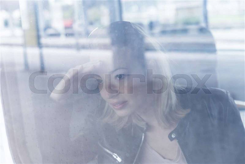Young blond woman traveling by train, looking out window while sitting in train. Through the window shot with reflections of exterior in glass, stock photo