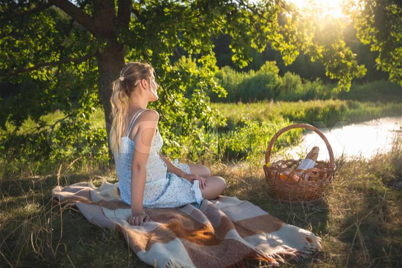 Toned image of beautiful young woman having picnic under big tree looking at evening sun over lake, stock photo