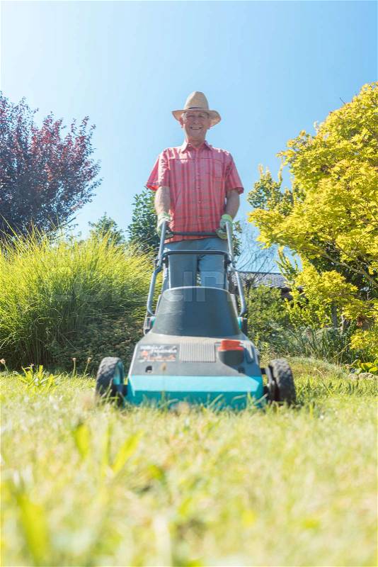 Low-angle view of an active senior man smiling and looking at camera while using a grass cutting machine in the garden, stock photo