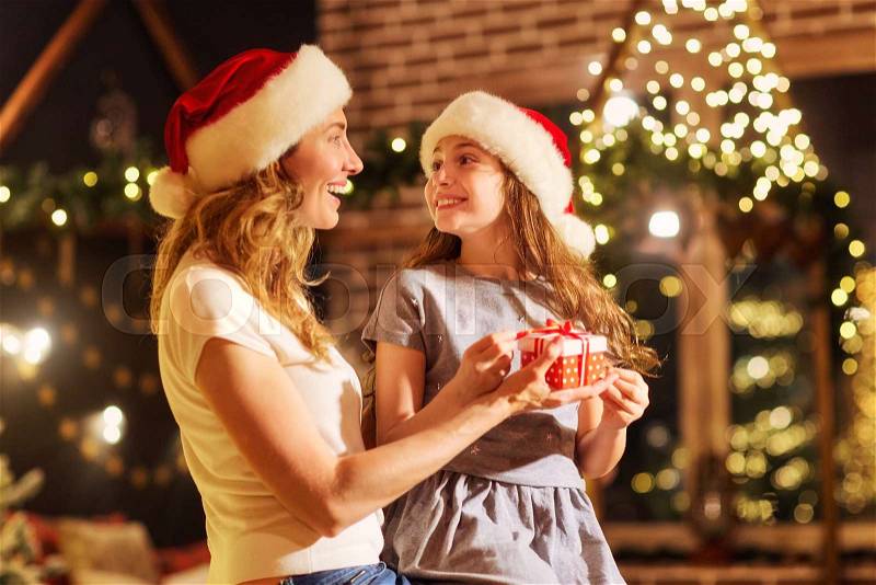 Mother and daughter in Santa hats give presents in a room with a fireplace and a Christmas tree on Christmas, stock photo