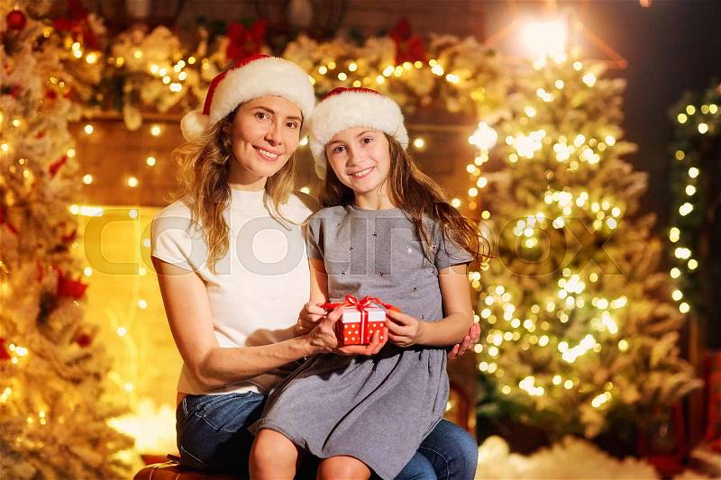 Mother and daughter in Santa hats give presents in a room with a fireplace and a Christmas tree on Christmas, stock photo