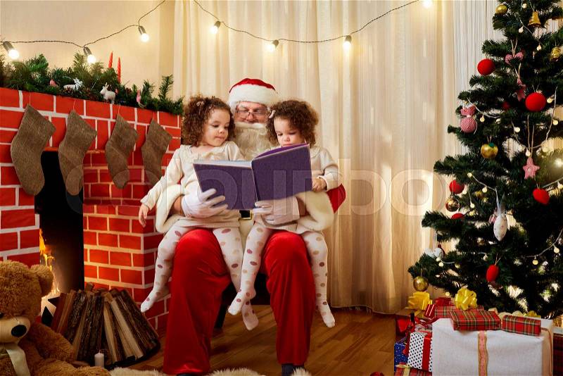 Santa Claus with children twin girls on his lap chiteyut book at Christmas, New Year in the room by the fireplace and Christmas tree. The concept of Merry Christmas, New Year, stock photo