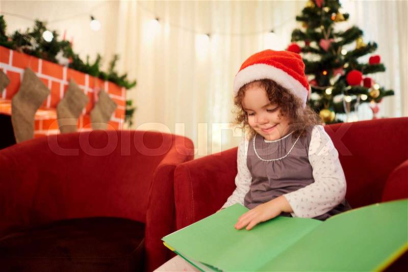The child, a little girl in the hat of Santa Claus reading a book sitting in a chair in a room with decorations, Christmas tree, fireplace in the new year, stock photo
