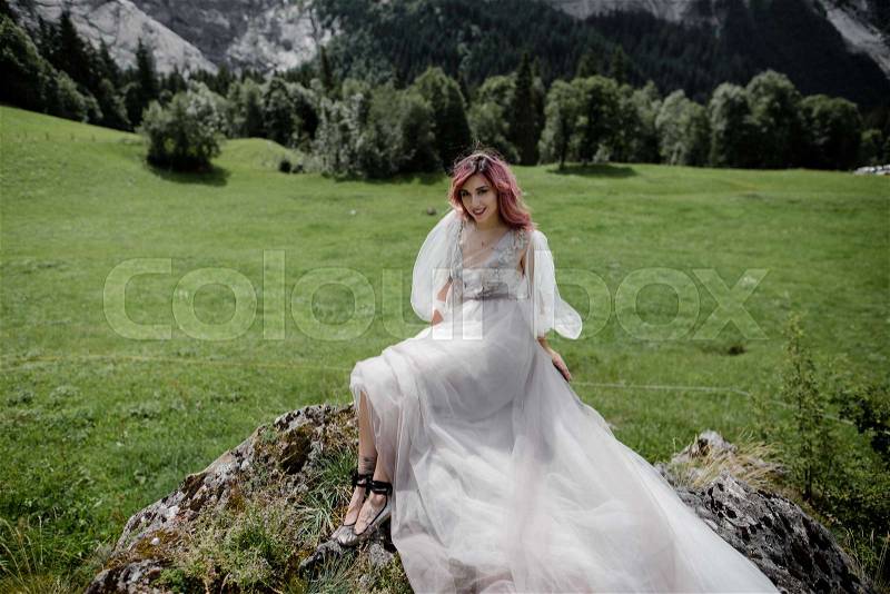 Beautiful young woman in wedding dress sitting on rock and looking at camera in mountain valley, Alps, stock photo