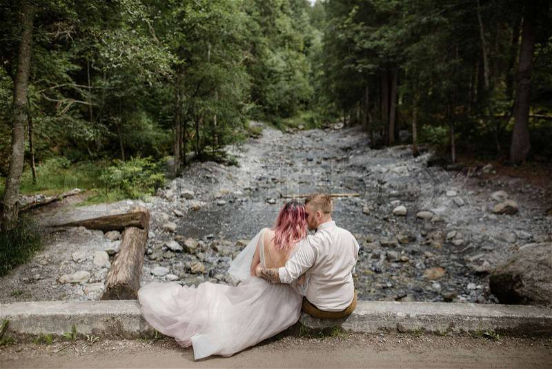 Back view of beautiful young bride and groom sitting on bridge above mountain river in Alps, stock photo
