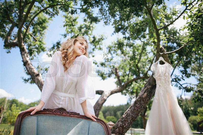 Low angle view of happy young bride in white robe leaning at chair while wedding dress hanging on tree, stock photo