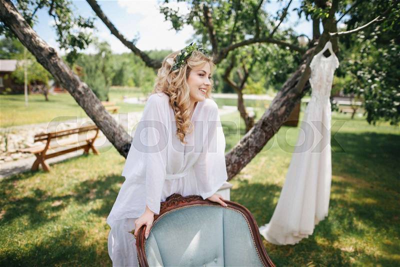 Beautiful smiling young bride in floral wreath and white robe leaning at chair and looking away outdoors, stock photo