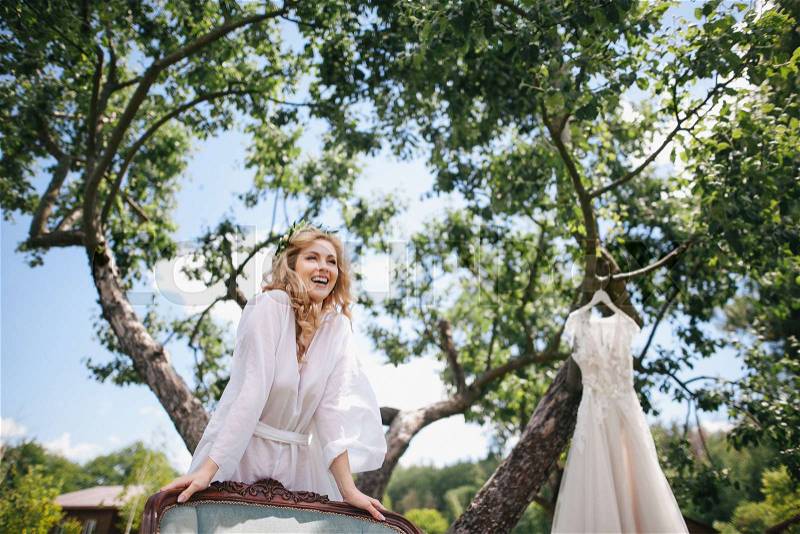 Happy young bride in white robe leaning at chair while wedding dress hanging on tree, stock photo