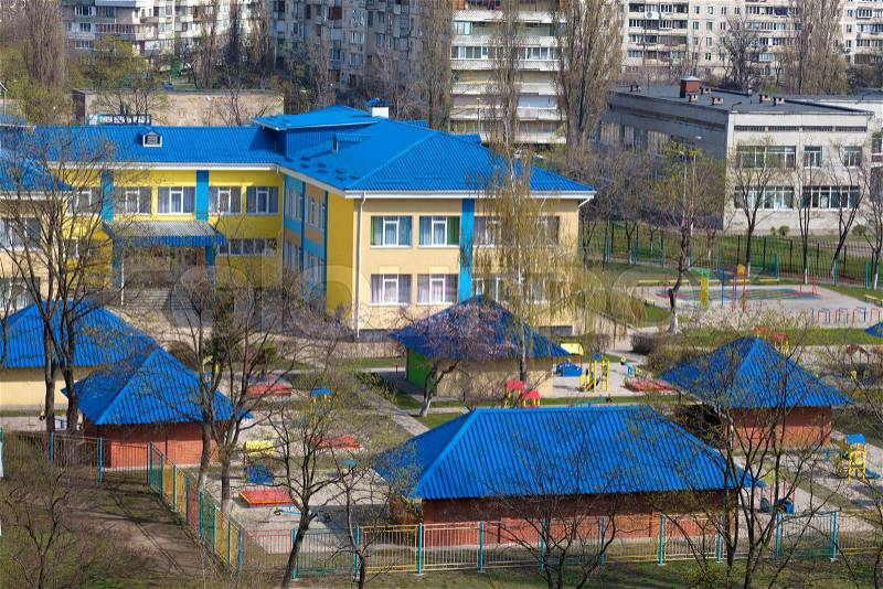 The bright blue roof of the kindergarten is illuminated by the spring sun against the background of gray urban high-rise buildings, stock photo