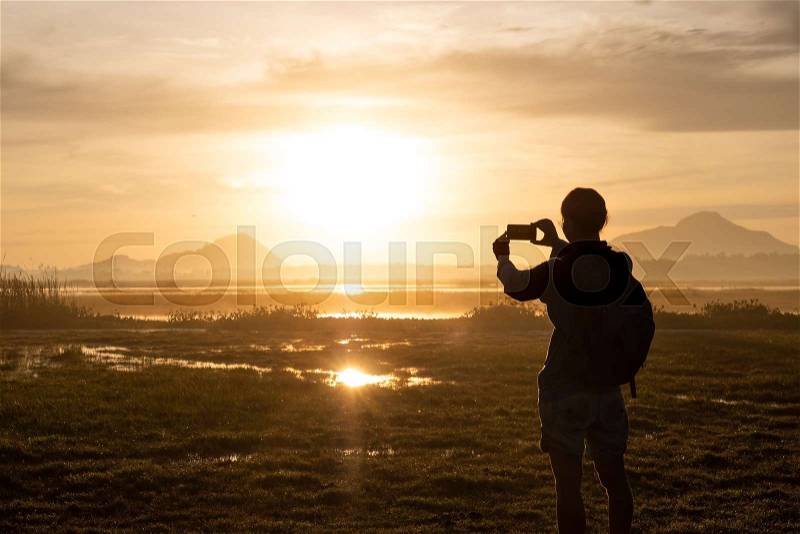 Silhouette of a woman holding a smartphone taking pictures outside during sunrise or sunset. Travelling and vacation concept, stock photo