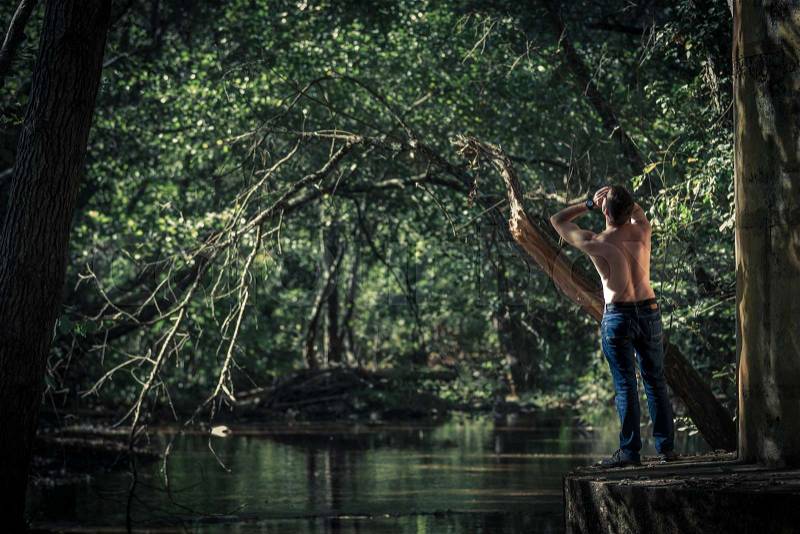 Back view of shirtless man in jeans standing and shouting on background of green pond and trees, stock photo