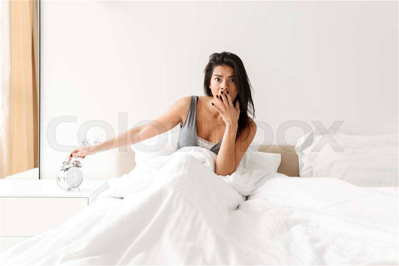 Image of disappointed woman 20s turning off ringing alarm clock on nightstand, while waking up and being late, stock photo