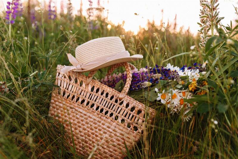 Vintage straw bag and hat, bouquet of wildflowers chamomile in high grass at sunset field, stock photo