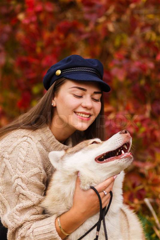 Portrait of young beautiful woman in hat sitting in autumn park hugging white husky dog. happiness and friendship. girl playing with pet on red trees background, stock photo