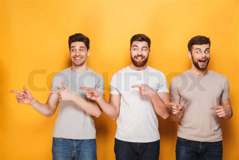 Three young excited men pointing away at copy space isolated over yellow background, stock photo