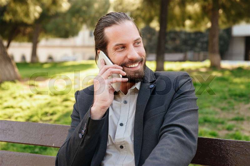 Photo of happy bearded man 30s in formal suit having mobile conversation, while sitting on bench in city park during sunny day, stock photo