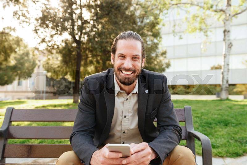 Picture of joyful handsome man with tied hair looking at camera and holding cell phone, while sitting on bench in city park during sunny day, stock photo