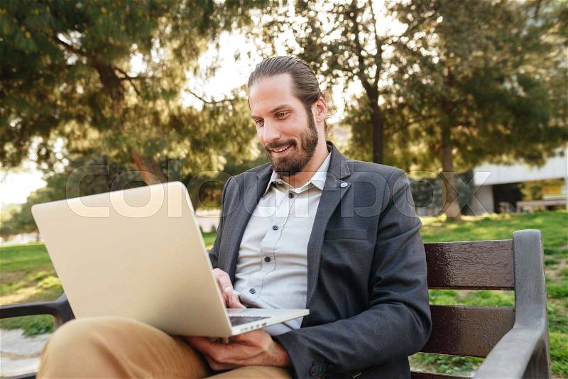 Picture of businesslike handsome man with tied hair working on silver laptop, while sitting on bench in city park during sunny day, stock photo