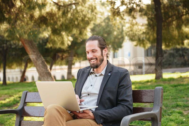 Photo of stylish adult man with tied hair laughing while working on silver laptop, and sitting on bench in green park, stock photo