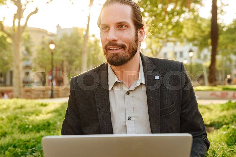 Picture closeup of businesslike handsome man with tied hair working on silver laptop, while resting in city park during sunny day, stock photo
