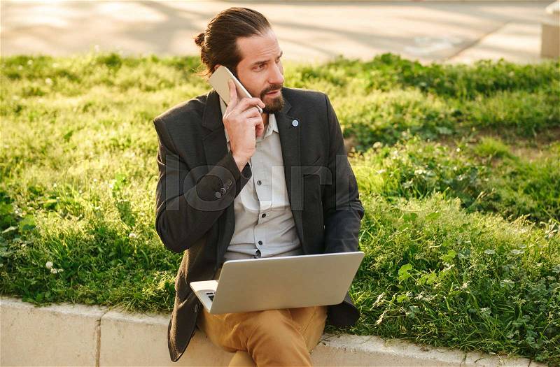 Photo closeup of caucasian business man with tied hair working on silver laptop and talking on mobile phone, while sitting in green park during sunny day, stock photo