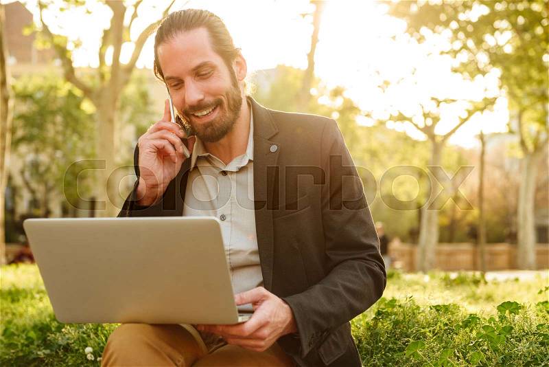 Picture closeup of working business man with tied hair looking at silver laptop and having mobile call, while sitting in park during sunny day, stock photo
