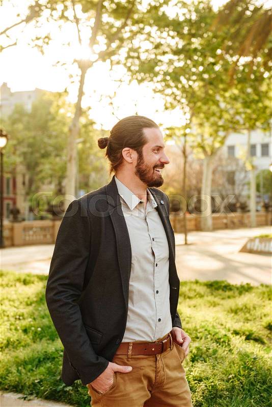 Profile picture of cheerful handsome man with tied hair smiling, and standing with hands in pockets in green park during sunny day, stock photo