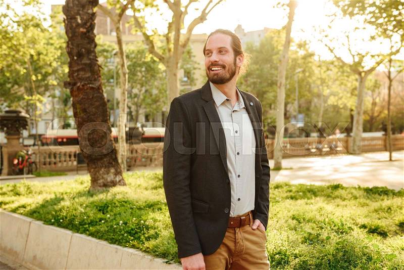 Portrait of positive handsome man 30s with tied hair smiling, and standing with hand in pocket in green park during sunny day, stock photo