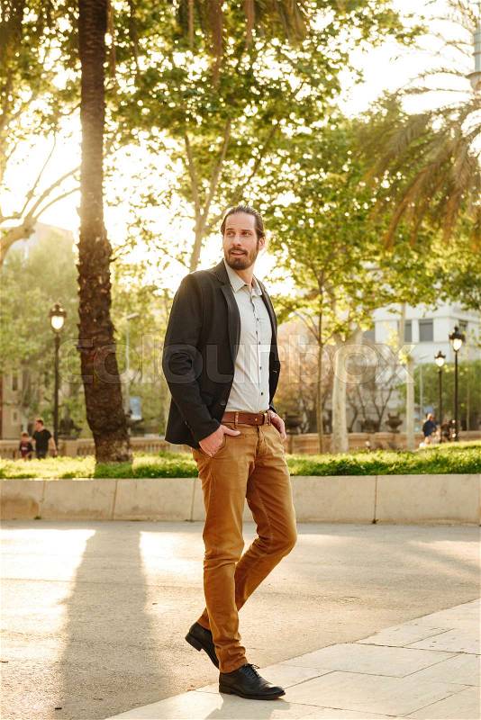 Full length image of unshaved attractive guy 30s with tied hair posing with hands in pockets, during walking in green park, stock photo